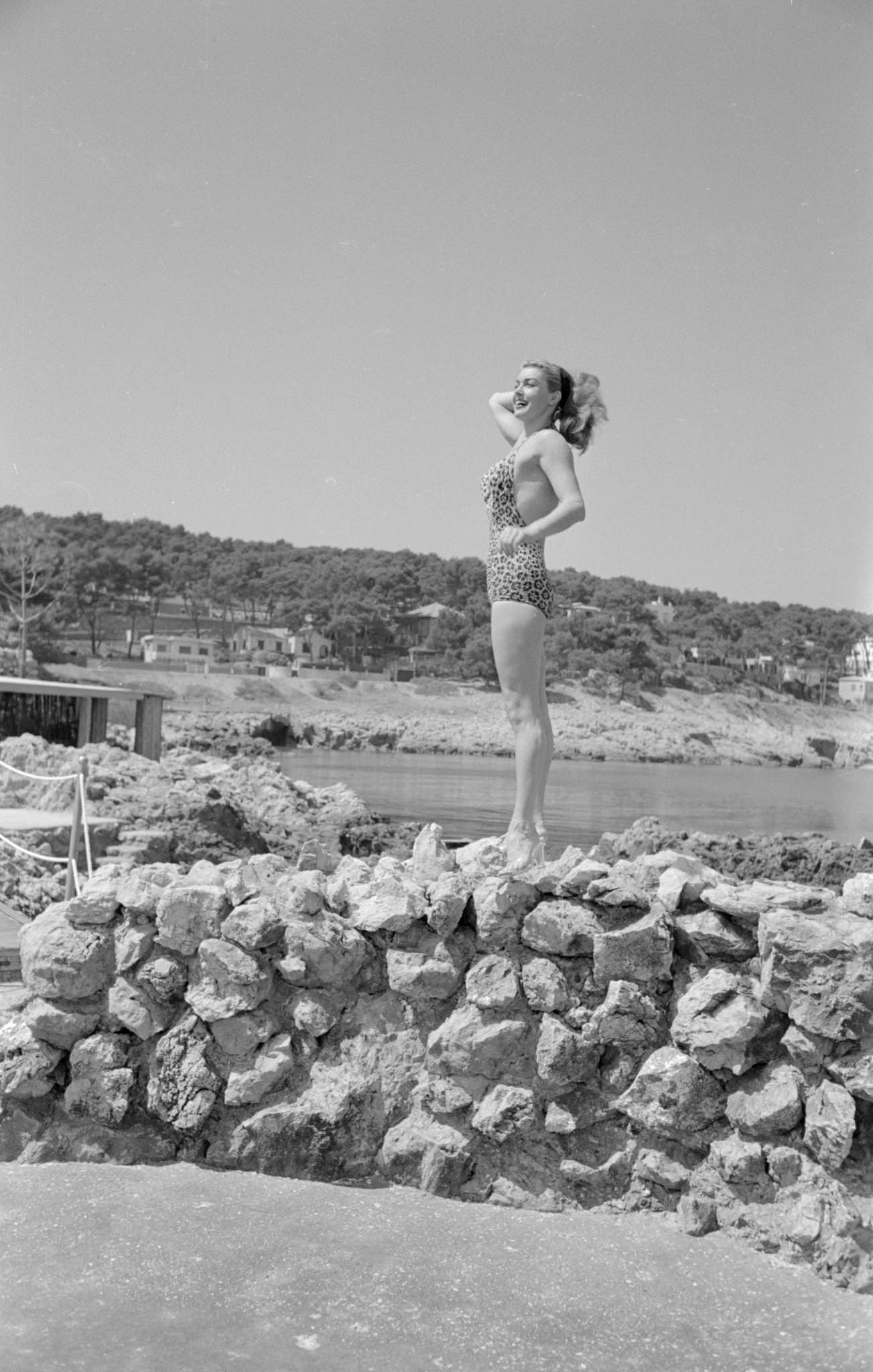 Esther Williams Smiling And Standing On A Rock In Clear Heels, Wearing A Leopard-Print Swimsuit, One Hand In Her Hair.