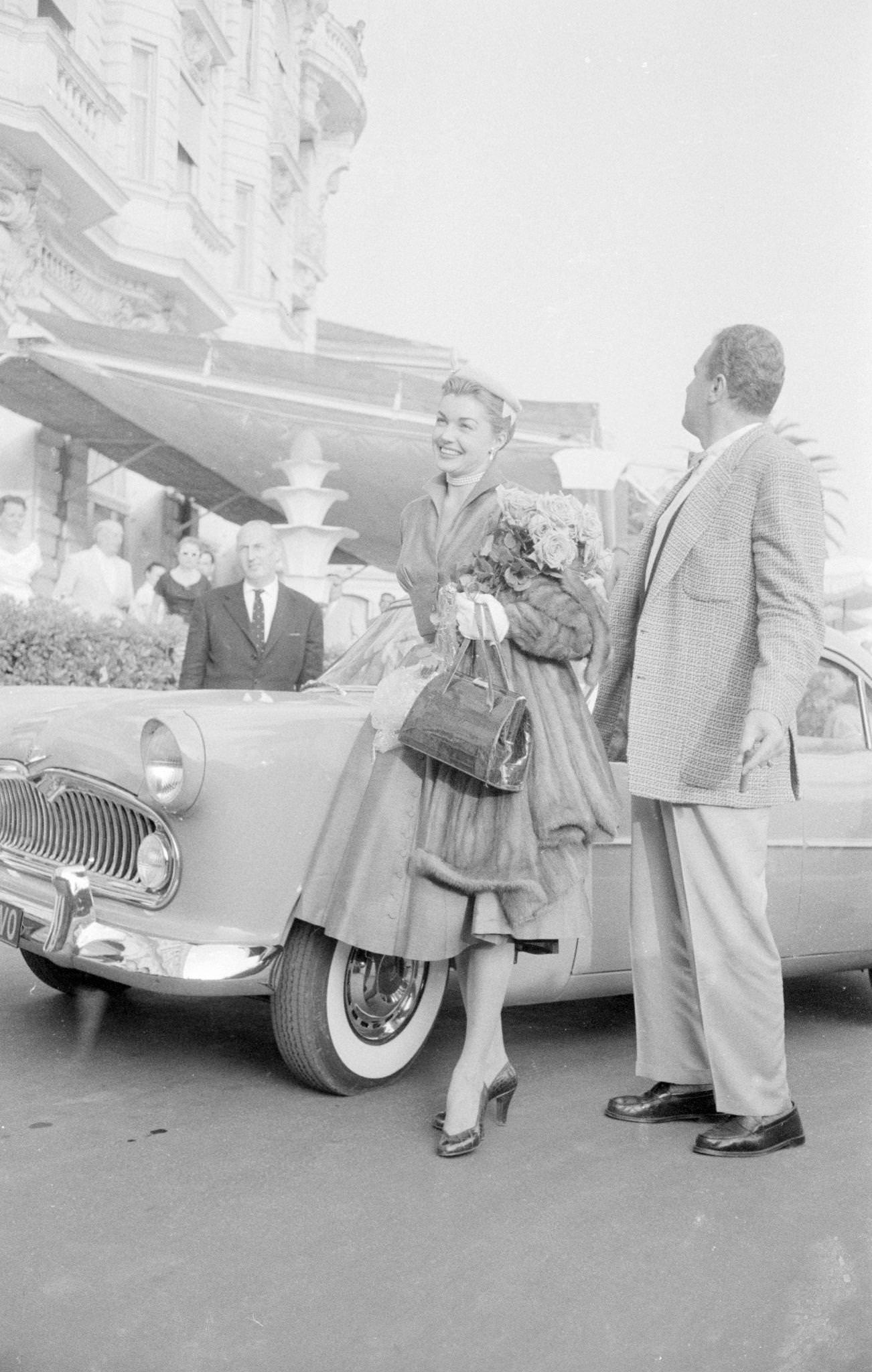 Esther Williams Holding A Bouquet Of Flowers, Wearing A Fur Coat, And Posing In Front Of A Parked Car Outside The Carlton Hotel