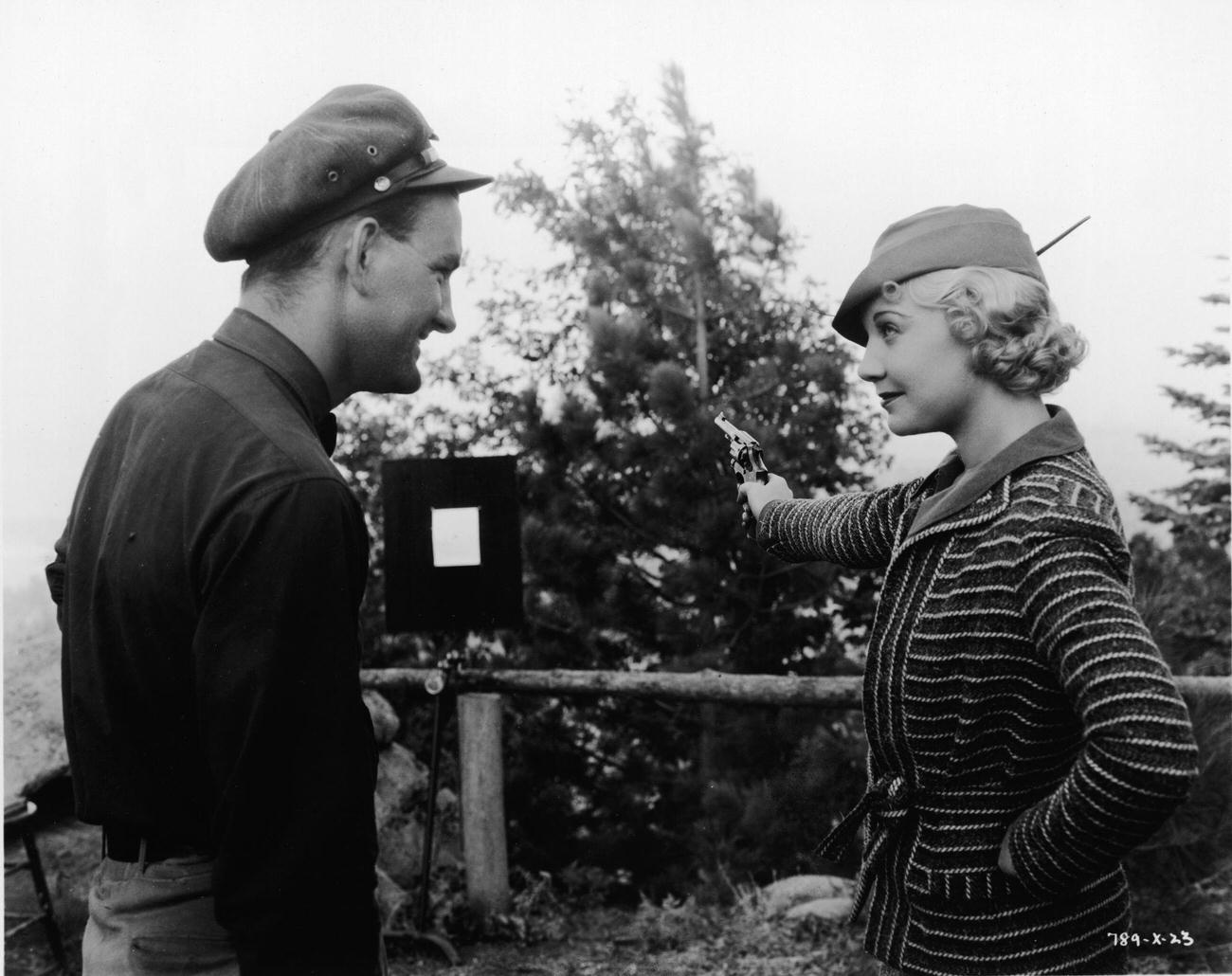 Una Merkel Rests One Hand On Her Hip And Points A Revolver At A Target In An Outdoor Shooting Range While Her Male Companion Stands By And Watches, 1930S.