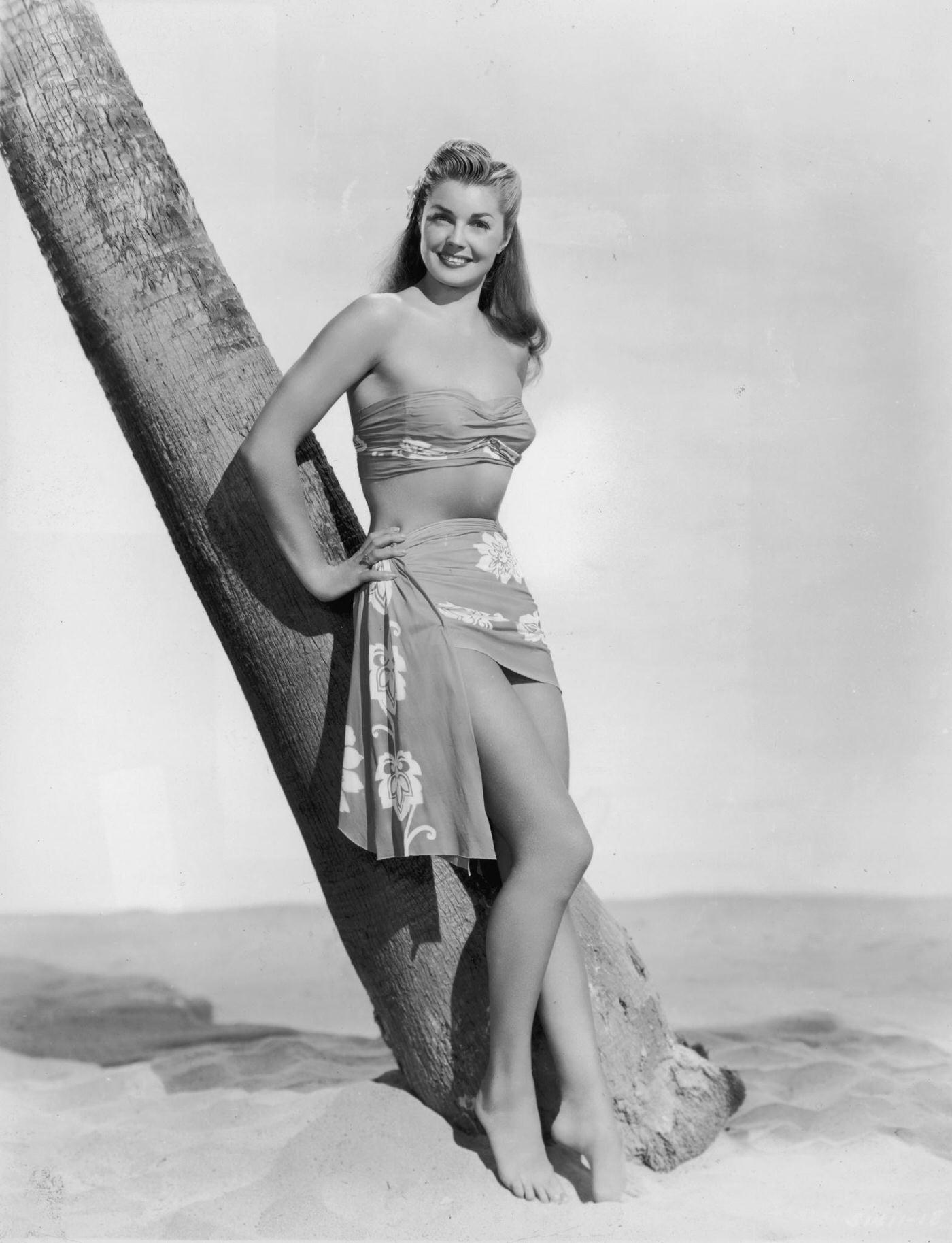 Esther Williams, Wearing A Bandeau Top And Sarong With A Tropical Print, Leaning Against A Tree Trunk In The Sand.