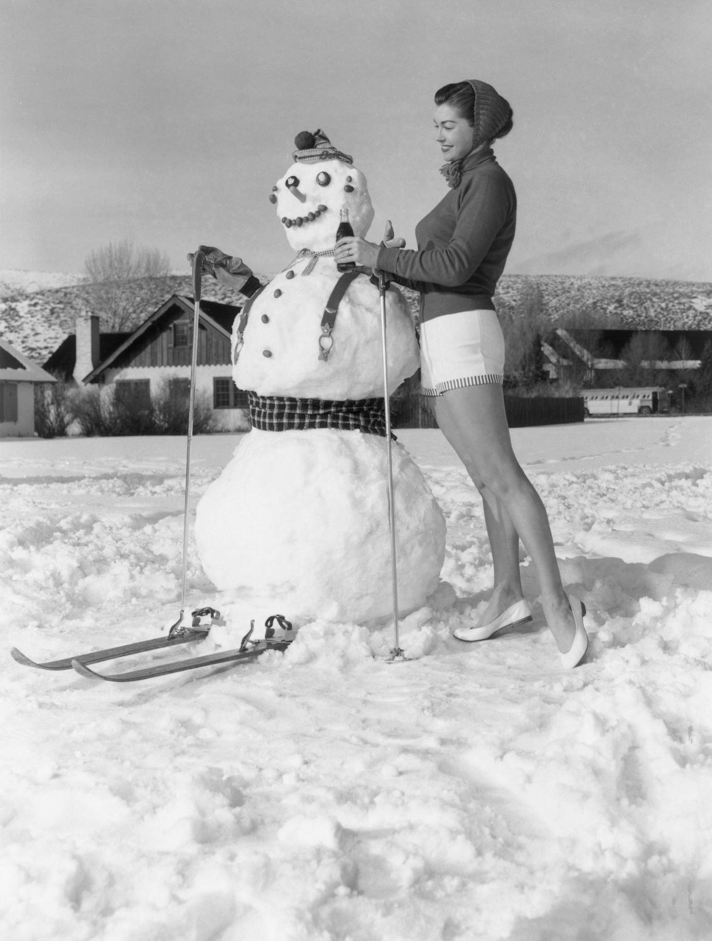 Esther Williams Offering A Bottle Of Coca-Cola To A Snowman On Skis On A Snow-Covered Yard, Sun Valley, Idaho. She Is Wearing Shorts And A Knit Scarf Over Her Hair.
