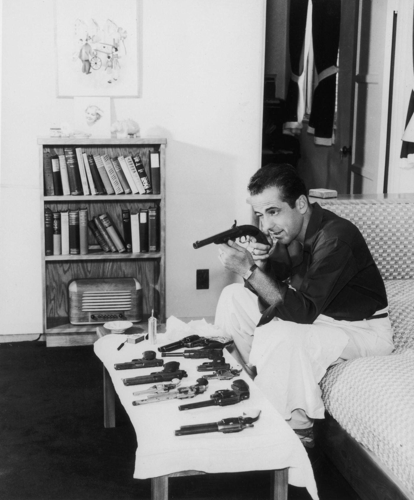 1939: Profile American Actor Humphrey Bogart , Seated On A Living Room Sofa Squinting As He Checks The Sights On A Gun He Holds In His Hands.
