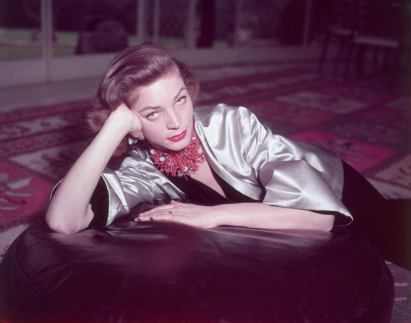 Lauren Bacall Wears Coral Necklace And Earrings With Gold Satin Jacket Over A Black Velvet Bodysuit While Seated On An Ottoman In 1955.