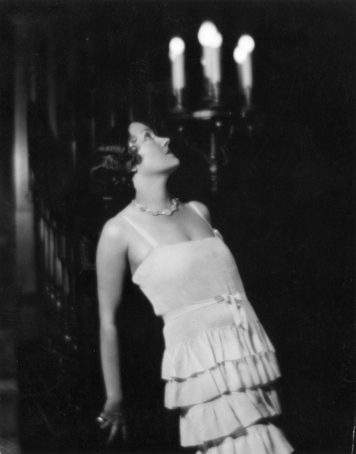 Gloria Swanson Poses By A Lit Candelabra, Wearing A Layered Gown With A Ribbon Belt, Circa 1930.