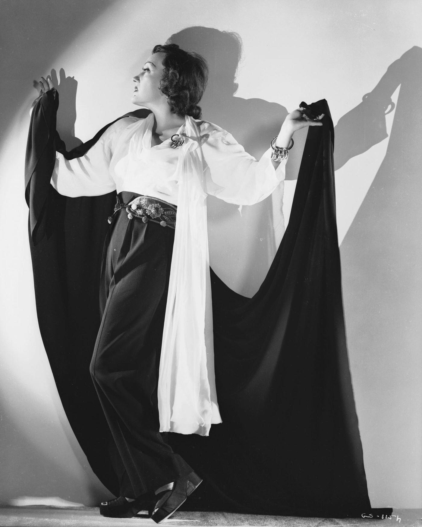 Gloria Swanson Poses In A Long Black Cloak, Lifting It Dramatically, Circa 1930.