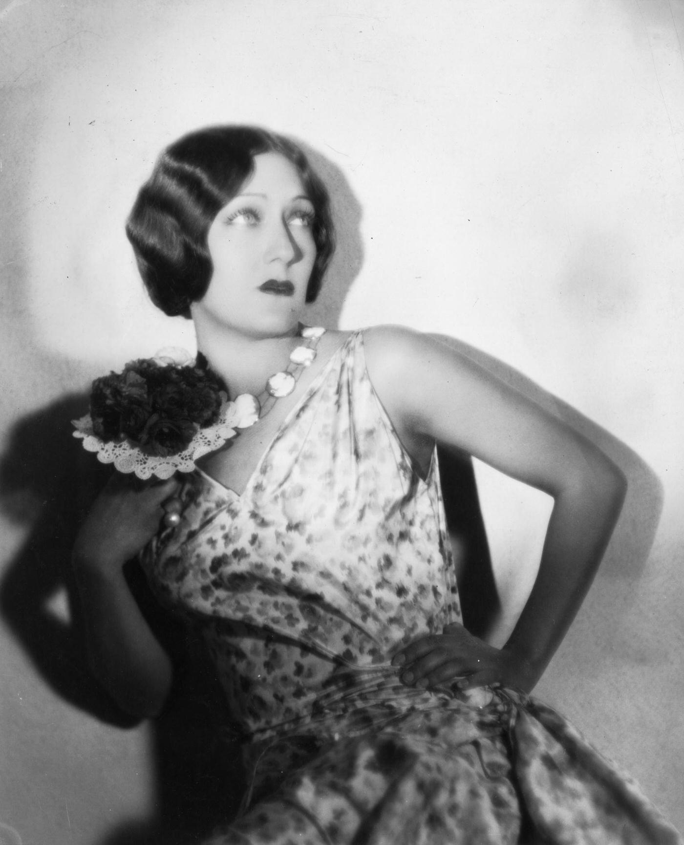 Gloria Swanson Wearing A Patterned Dress And Clutching A Posy Of Flowers, Circa 1930.