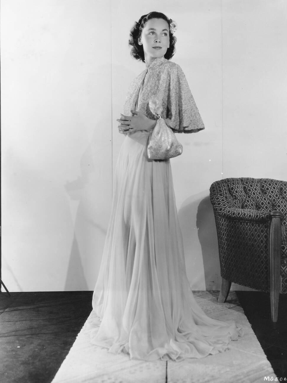 Maureen O'Sullivan Wears A Pale Pink Chiffon Gown With An Elbow-Length Cape. Her Silver Brocaded Bandana Bag Adds Formality. A Flower In Her Hair Complements The Look.