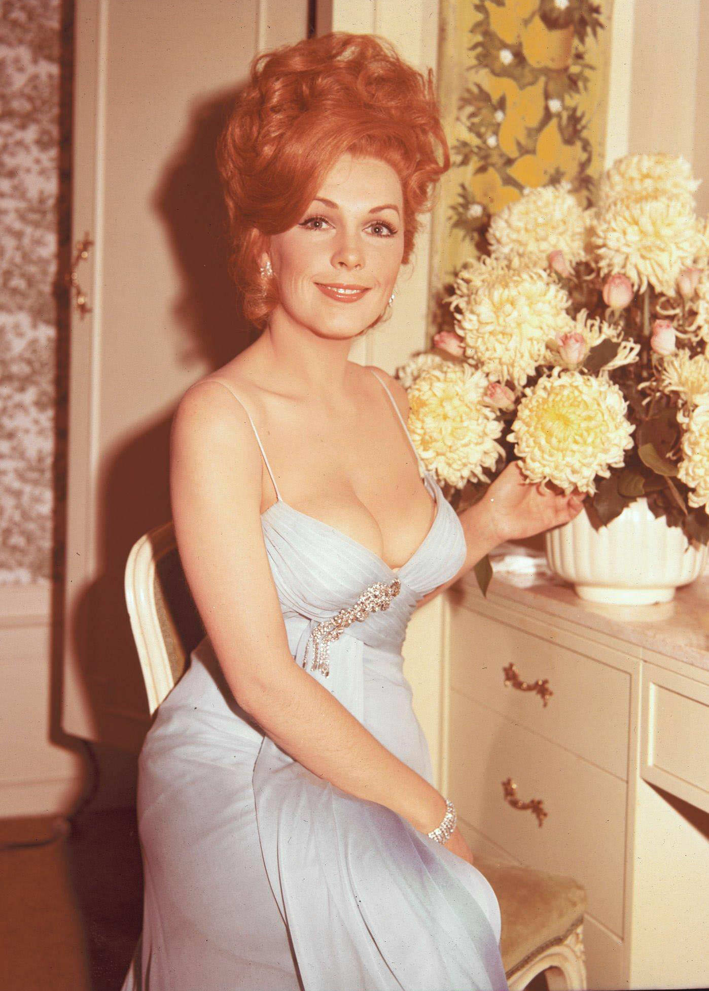 Stella Stevens Posing With Flowers, 1936. Seated In A Light Blue Low-Cut Dress, Pointing To A Vase Of Large Yellow Flowers.