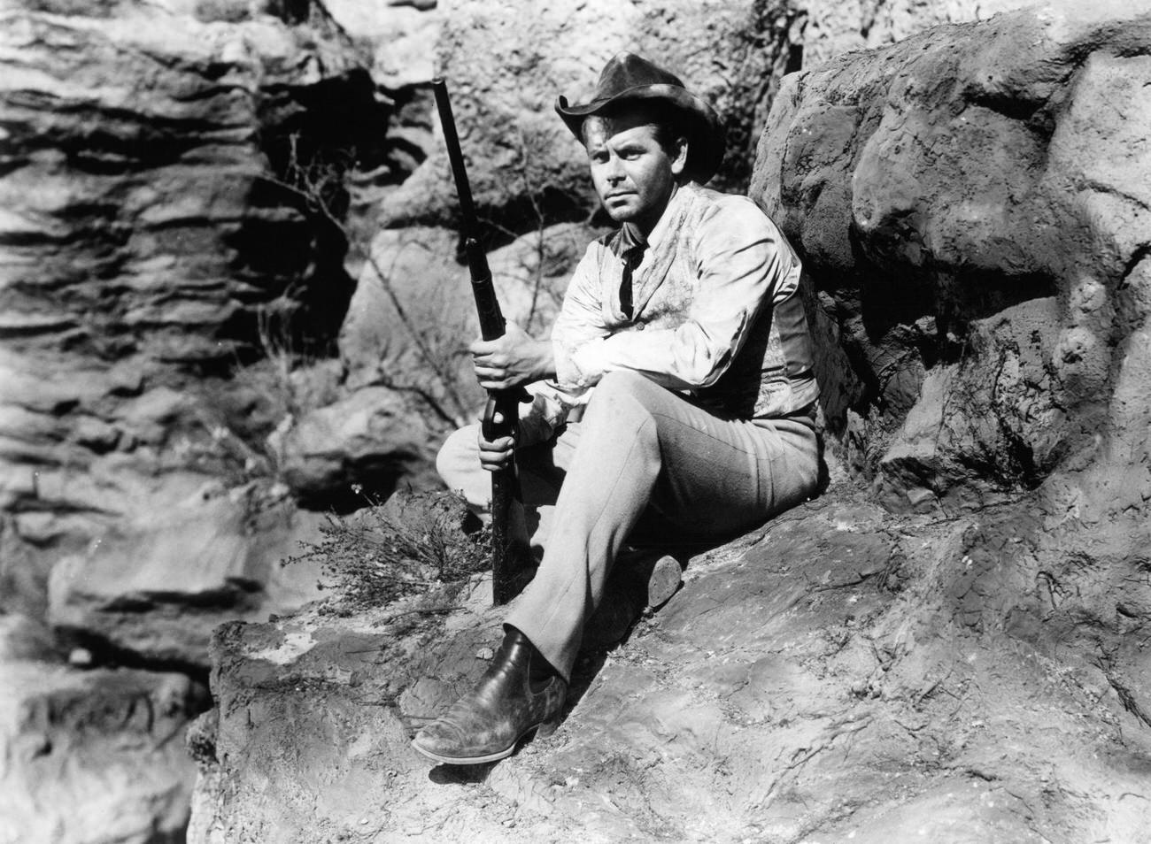 Glenn Ford Sitting On Rocks With A Rifle In 'Lust For Gold', 1949.