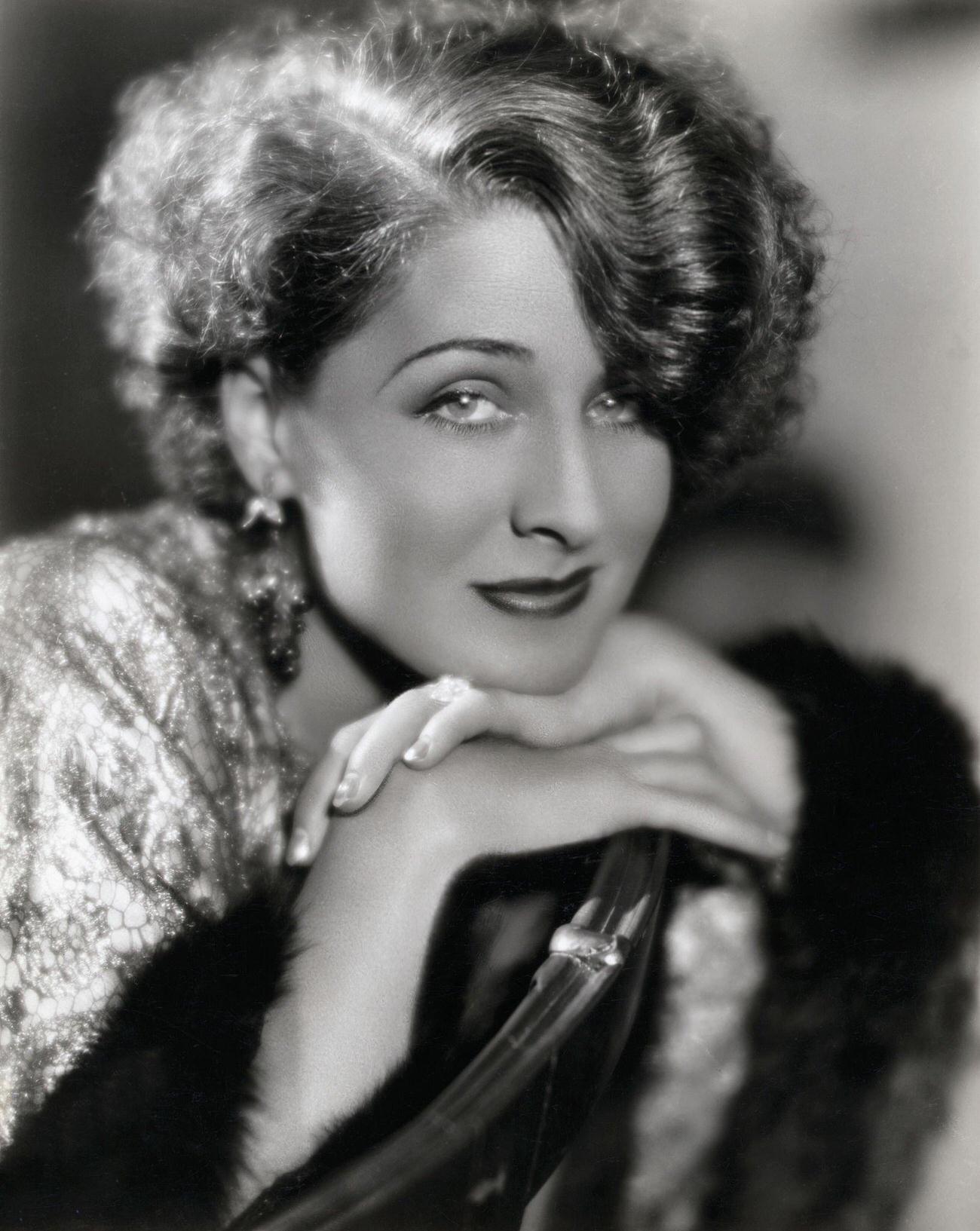 Norma Shearer Smiling With Her Chin On Her Folded Hands, Wearing A Shiny, Fur-Lined Stole.
