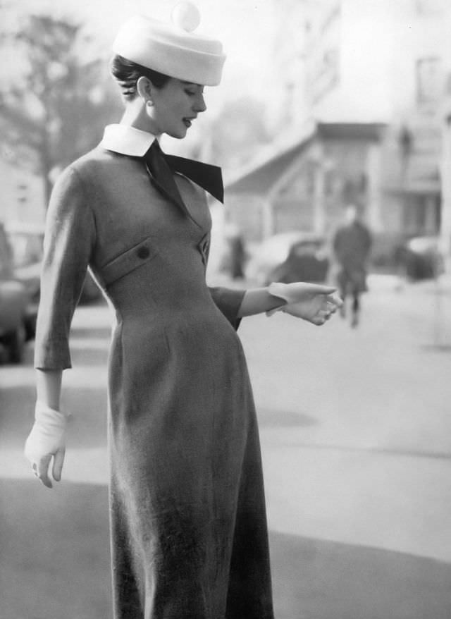 Model In Pewter Gray Tweed Dress Molded To The Waist, Has Half Belts Under The Bosom, Features A Starched Collar And Taffeta Tie, The White Hat Balances A Pom-Pom, By Jacques Heim, Photo By Richard Dormer, Harper'S Bazaar Uk, April 1956