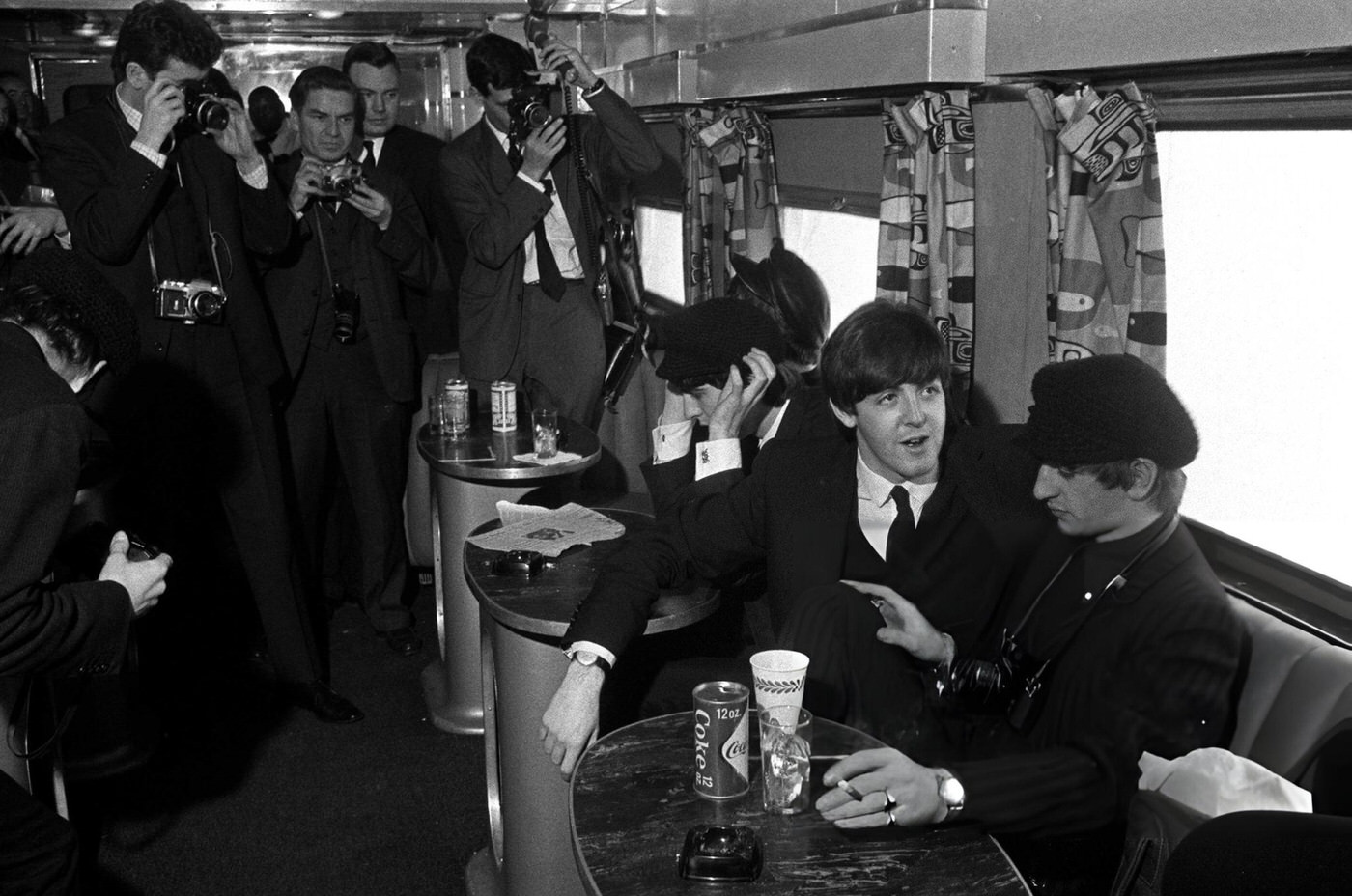 The Beatles Have Their Pictures Taken By A Group Of Photographers As They Sit At A Table With A Drink Of Coca Cola On The Train From New York To Washington During Their Tour Of The States, 1964.