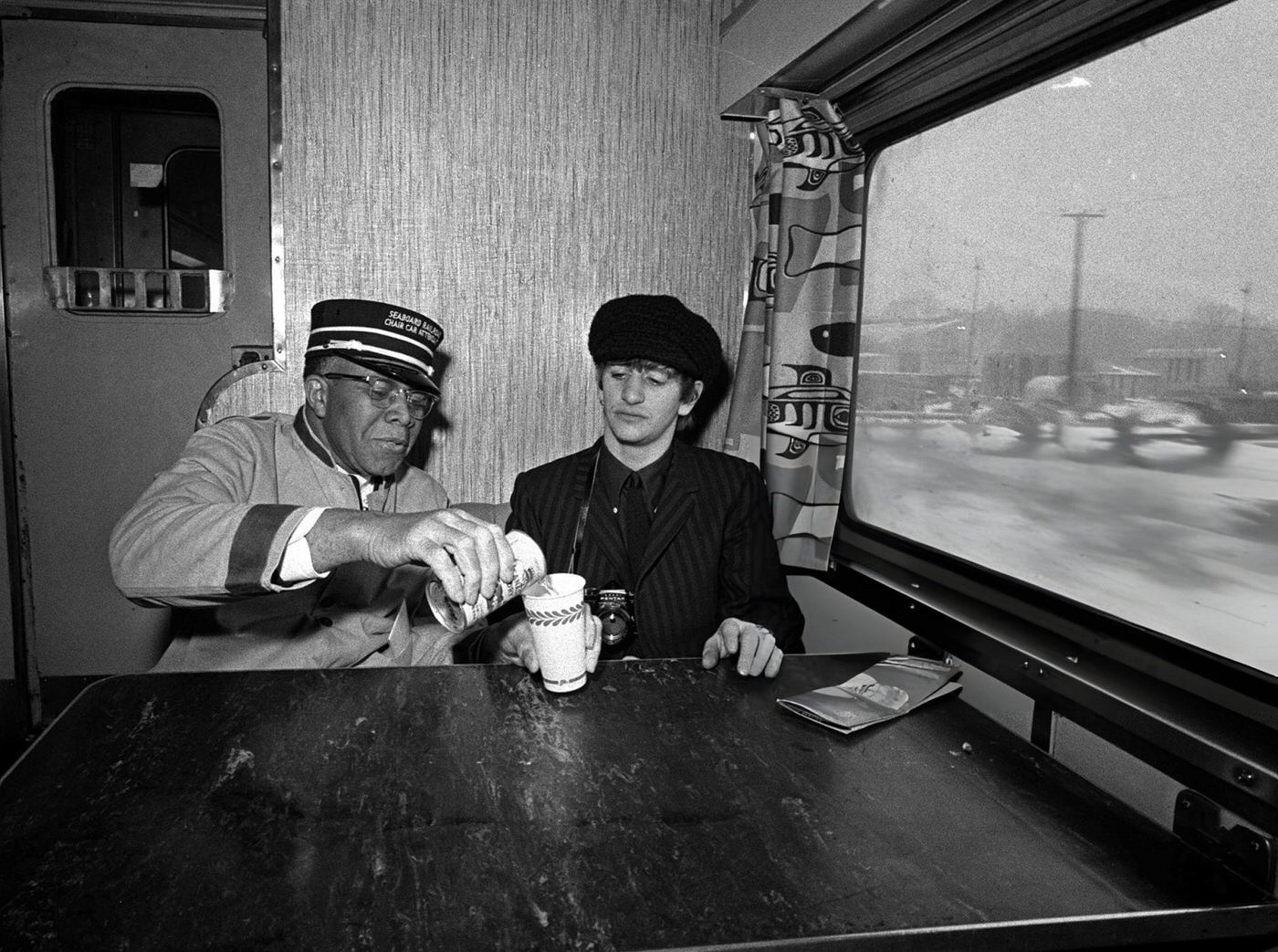 Ringo Starr, Drummer Of British Pop Group The Beatles Sitting At A Table As He Has A Drink Poured By Carriage Attendant John R. Ragsdale On Board The Train From New York To Washington During Their Tour Of The States, 1964.