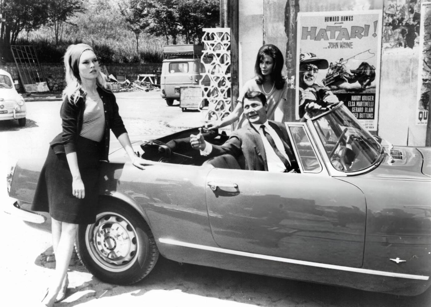 Brigitte Bardot Leaning Against A Car As Jack Palance Looks At Her In 'Contempt', 1963.