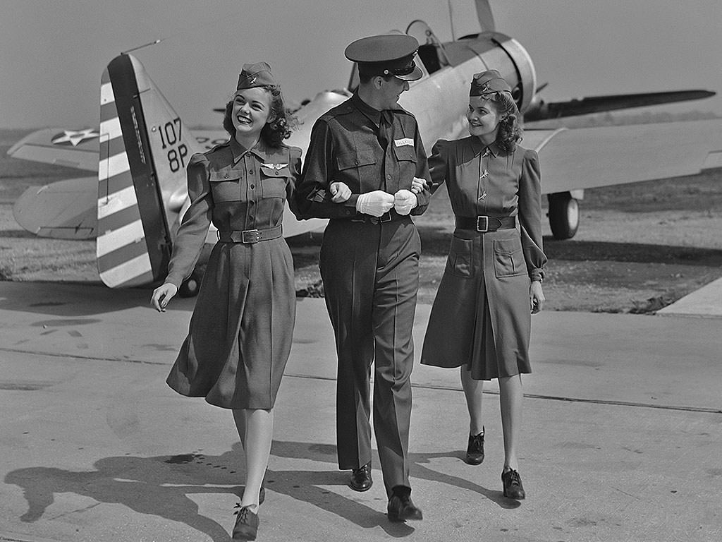 A Male Flight Attendant Walks With His Arms Linked With Two Female Flight Attendants In Front Of A Small Plane, 1940S.