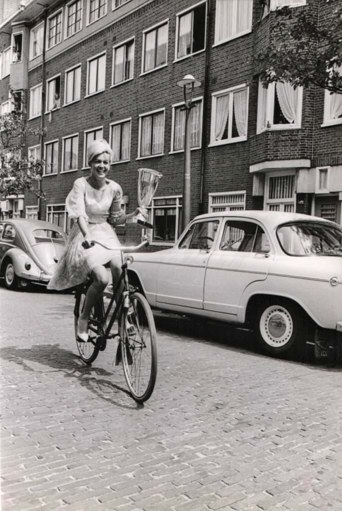 Elly Koot Rides A Bike, Wields The 1964 Miss Europe Trophy.